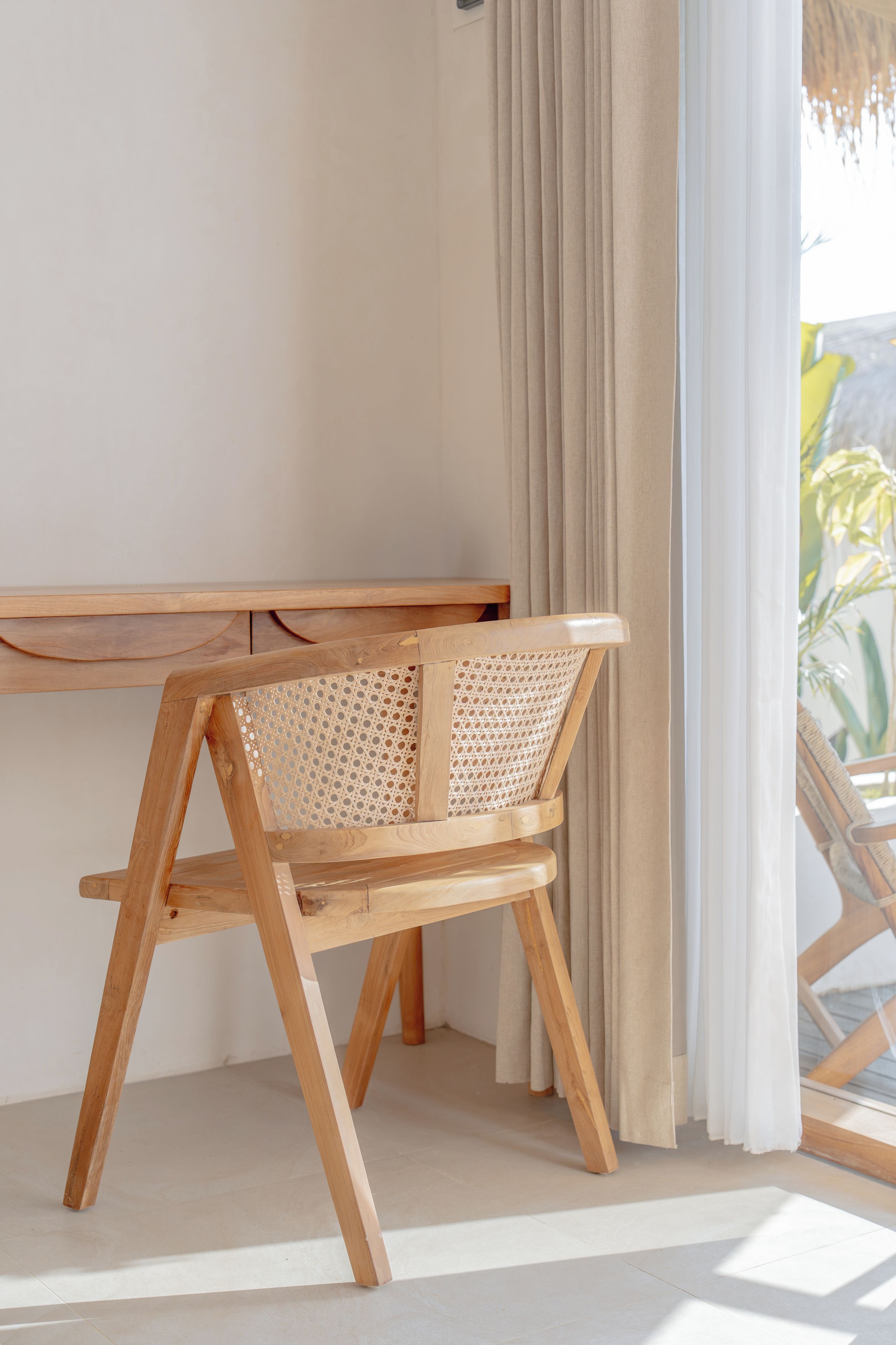 Een houten bureau met een geweven stoel, een raam met gordijnen en uitzicht op groen.