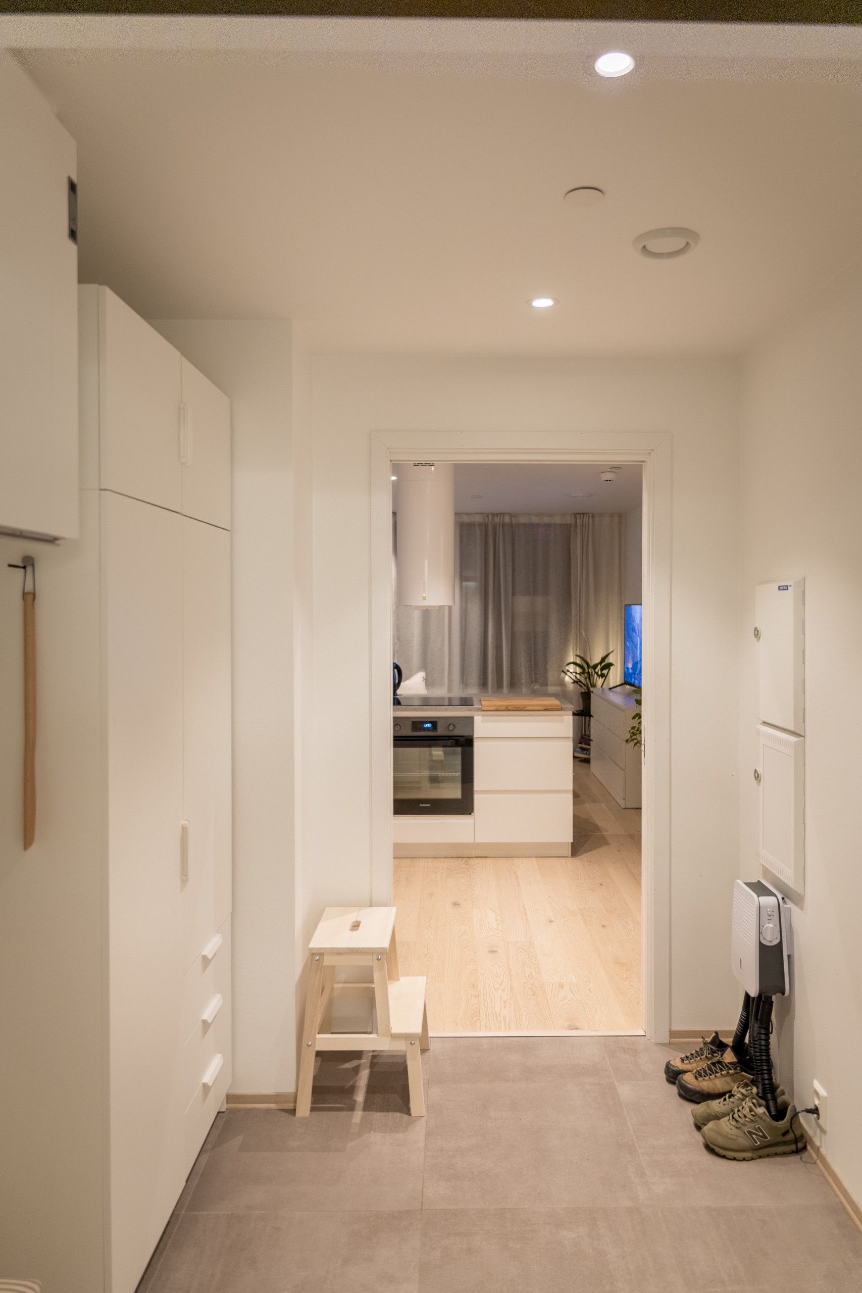 Un couloir étroit, avec une cuisine visible à travers une porte ouverte, un petit tabouret en bois et une paire de chaussures près de la porte.
