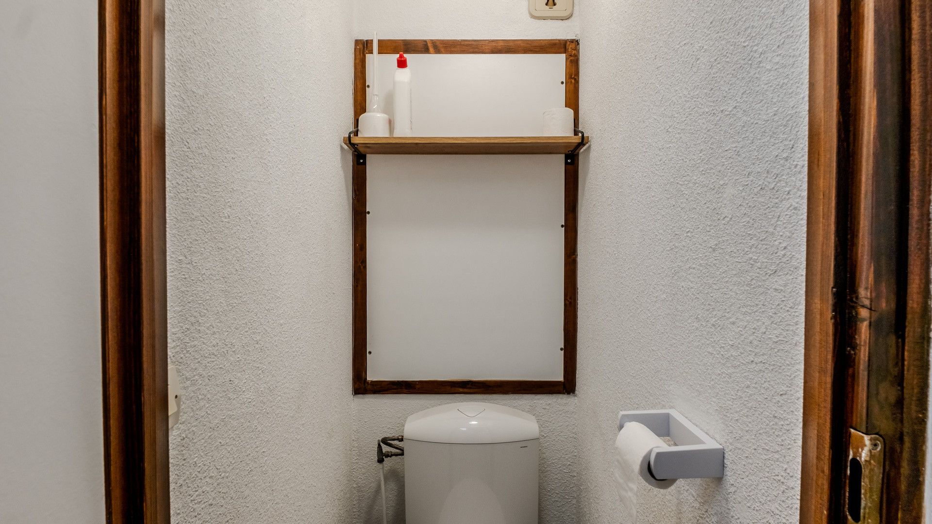 A bathroom with a toilet, a white tank, and a wooden shelf above it.