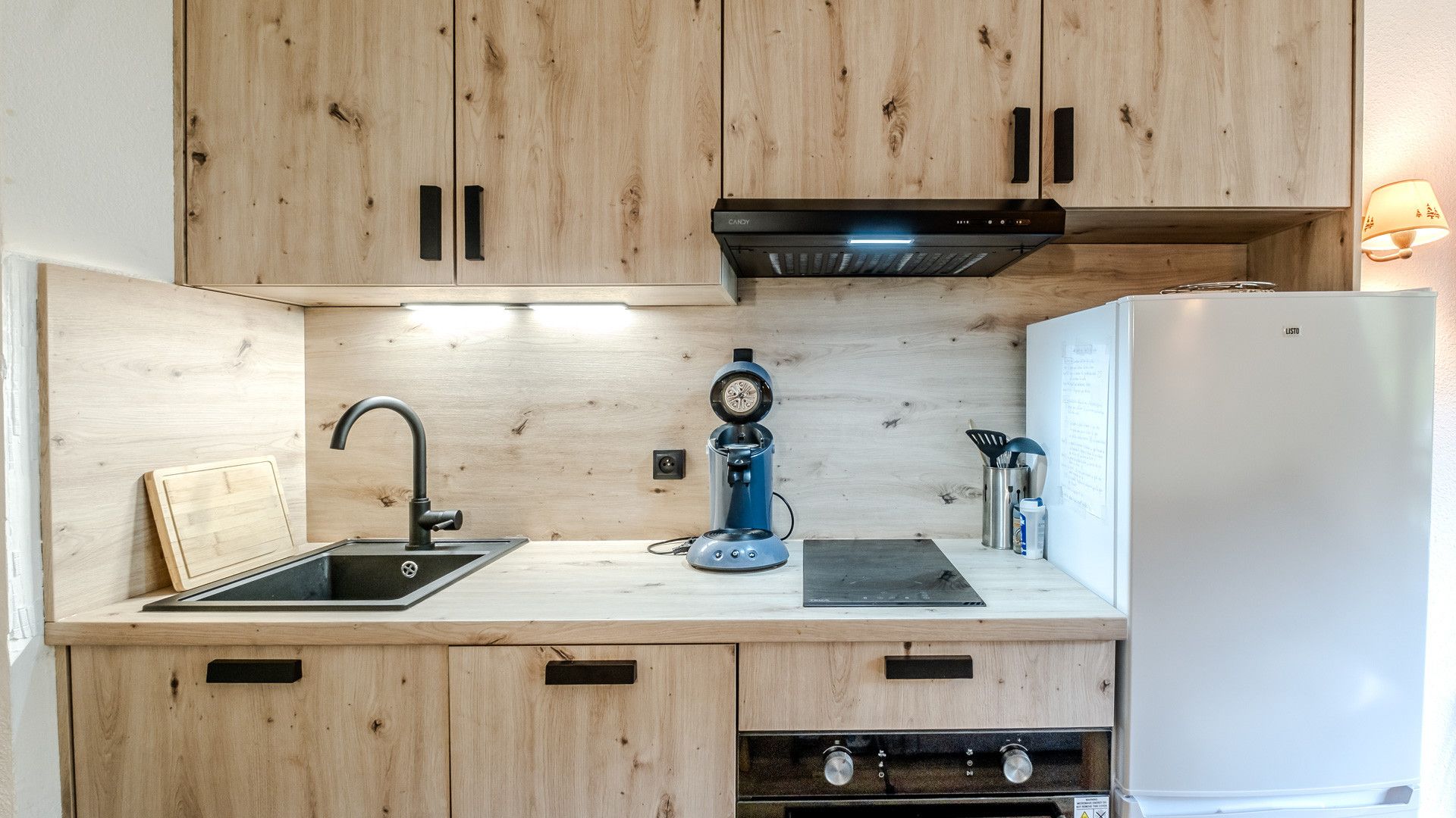 A compact kitchen with wooden cabinets, a sink, a coffee machine, a stove, and a refrigerator.