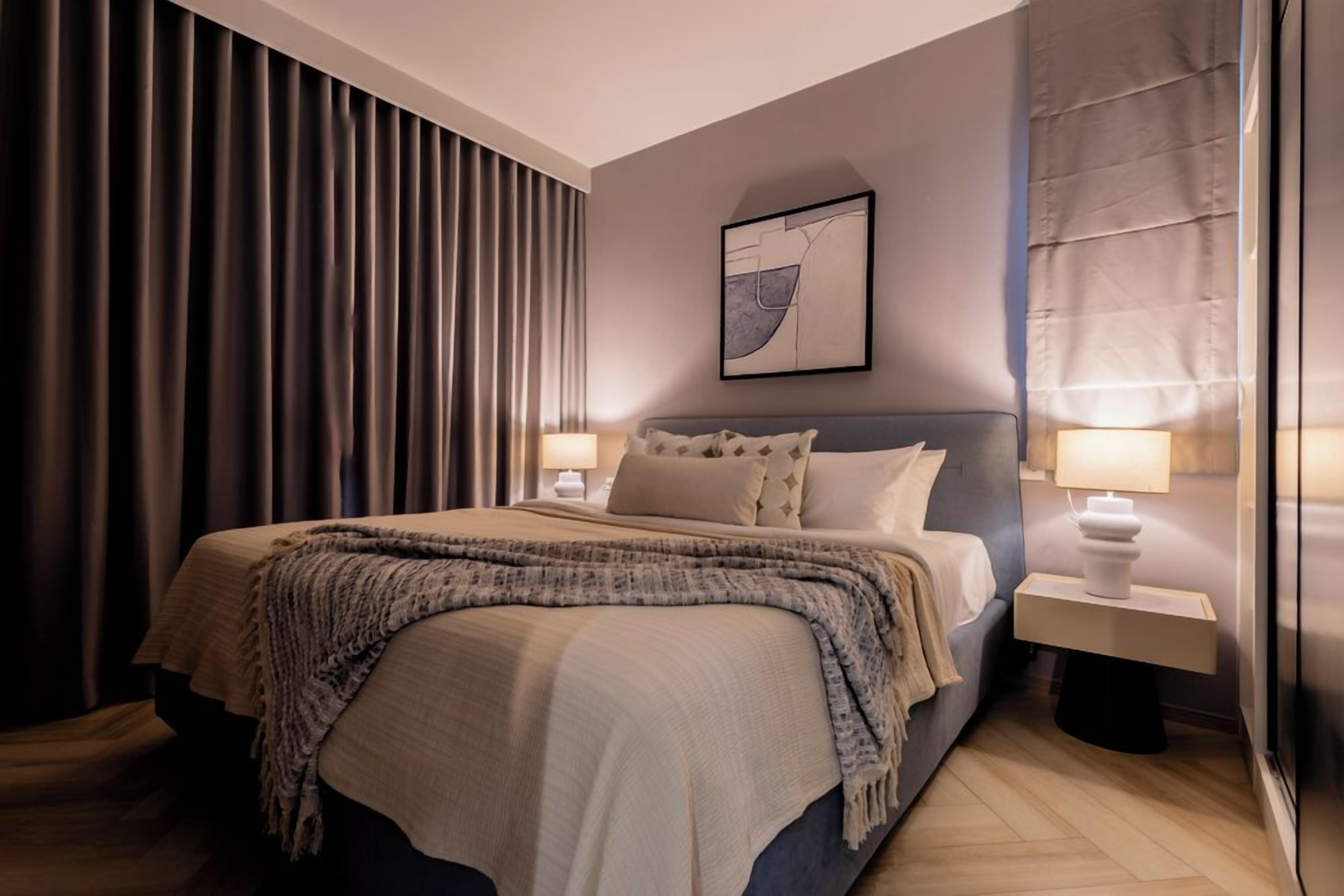 A modern hotel room with a large bed, two bedside tables with lamps, a wall-mounted artwork, and a large window with curtains.
