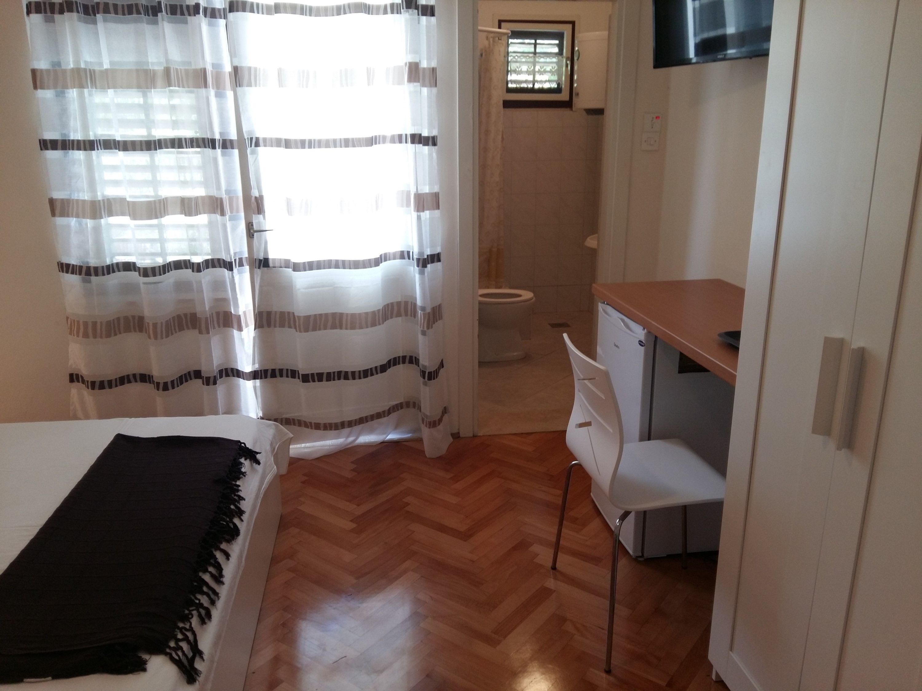 A bedroom with a bed, a desk, and a chair. There is a window with curtains, a toilet, and a TV mounted on the wall.