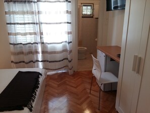 A bedroom with a bed, a desk, and a chair. There is a window with curtains, a toilet, and a TV mounted on the wall. - Spacious room near beach, with private bathroom & patio (Arbanija)