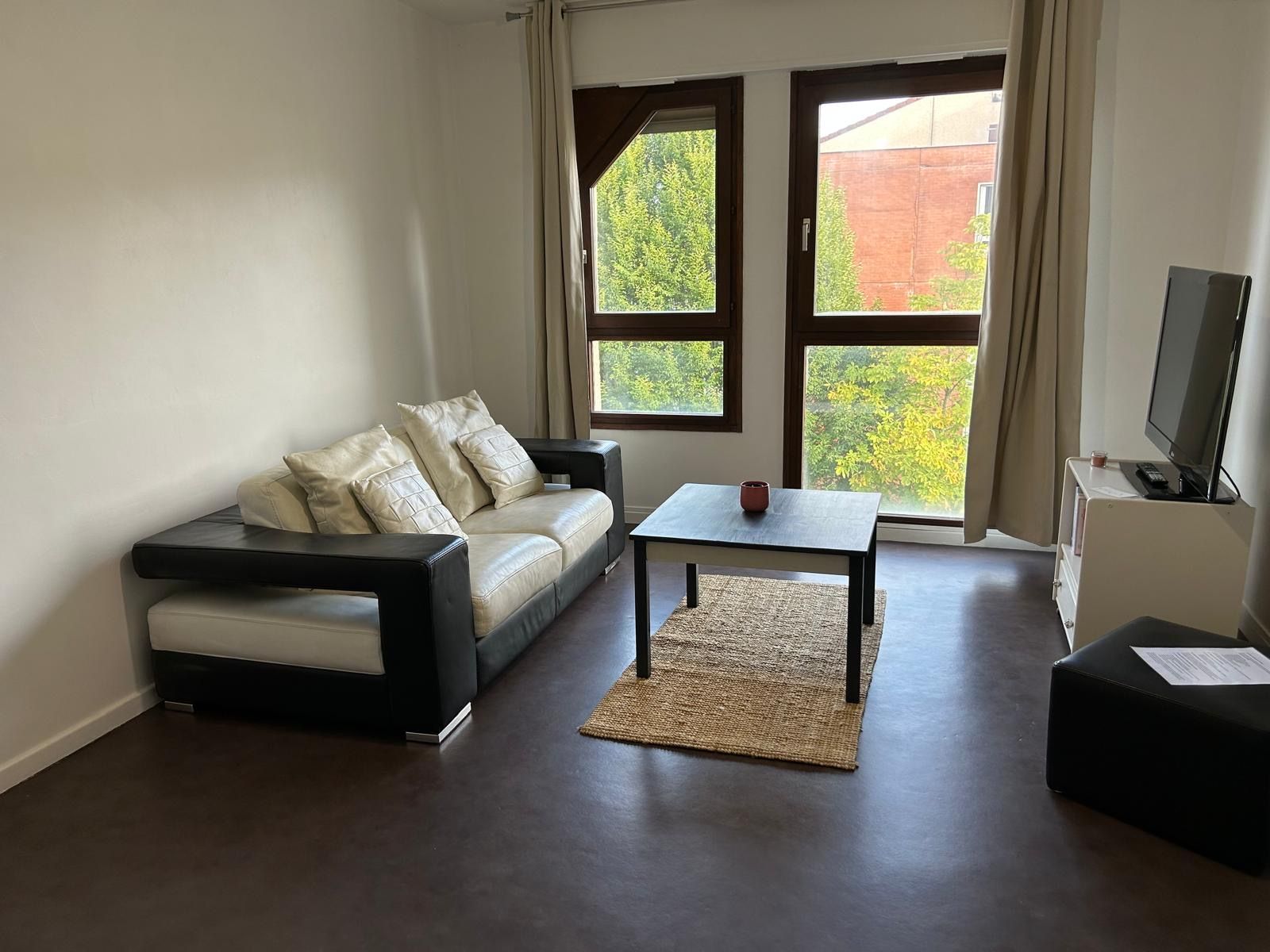 A living room with a sofa, a coffee table, a television, and large windows with a view of greenery.