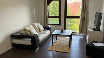 A living room with a sofa, a coffee table, a television, and large windows with a view of greenery.