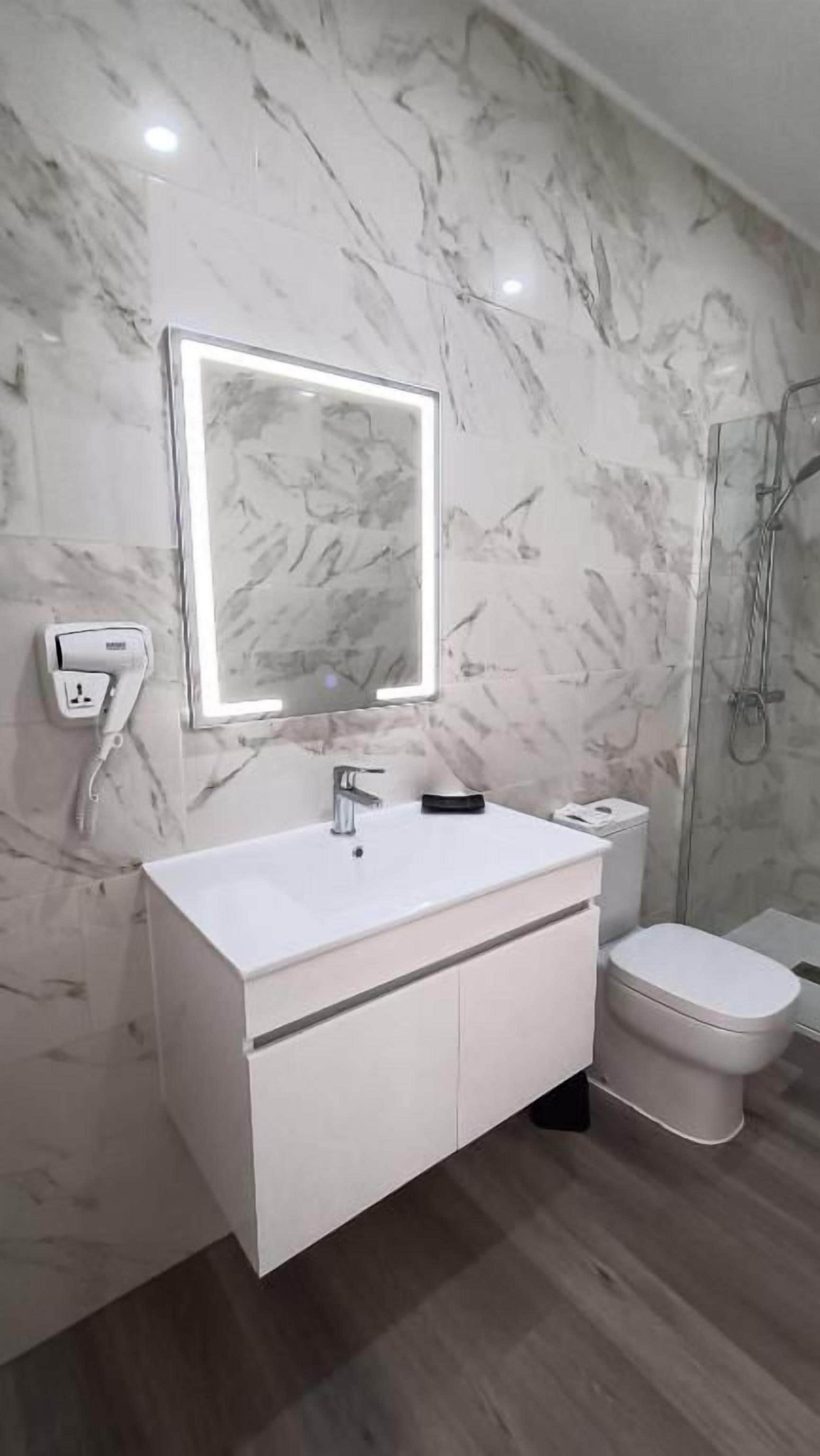 A bathroom with a marble wall, a white vanity, a mirror with lighting, a toilet, and a shower area.