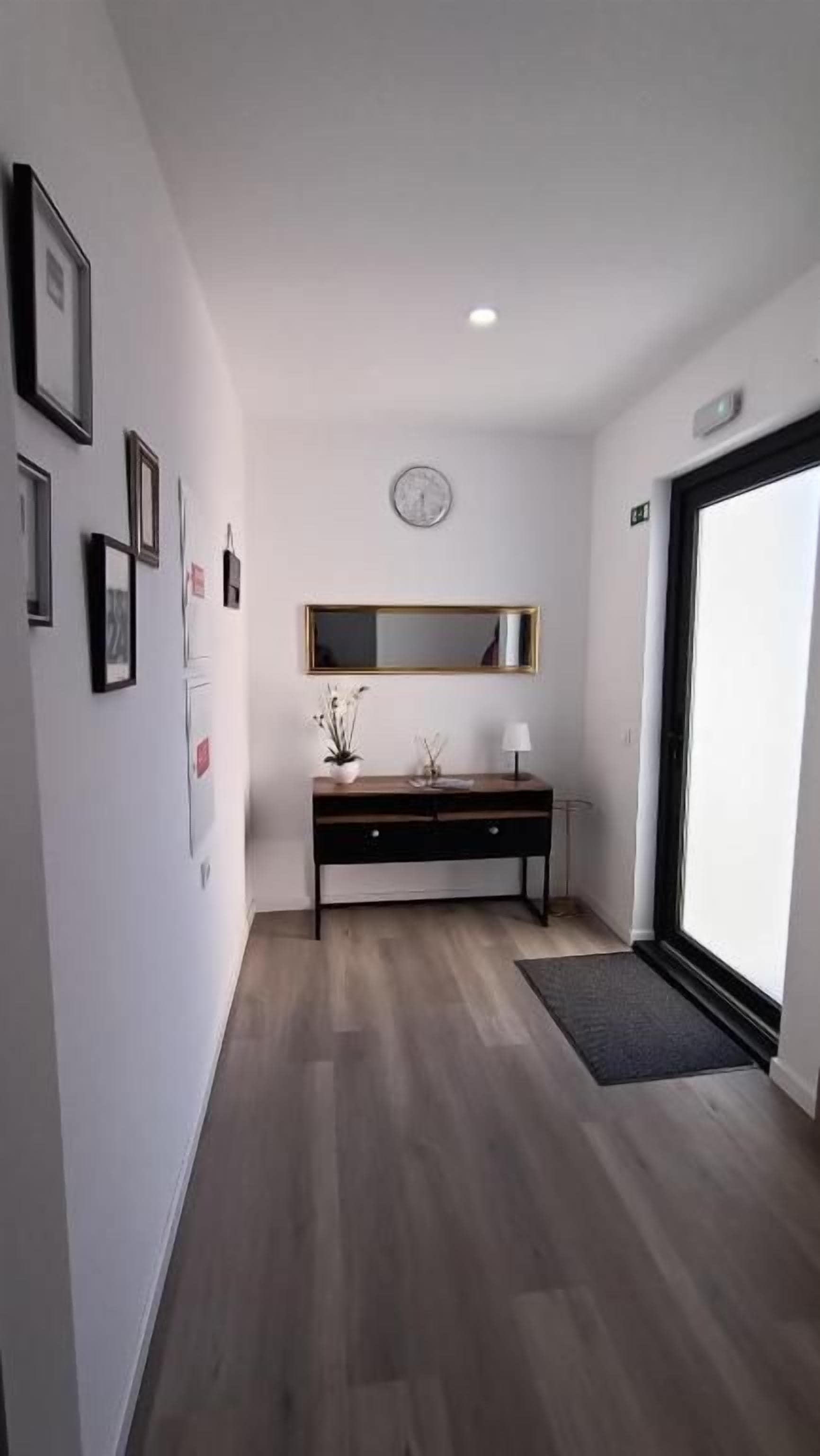 Un pasillo con piso de madera, una consola negra con espejo, cuadros en la pared y una puerta que da a otra habitación.