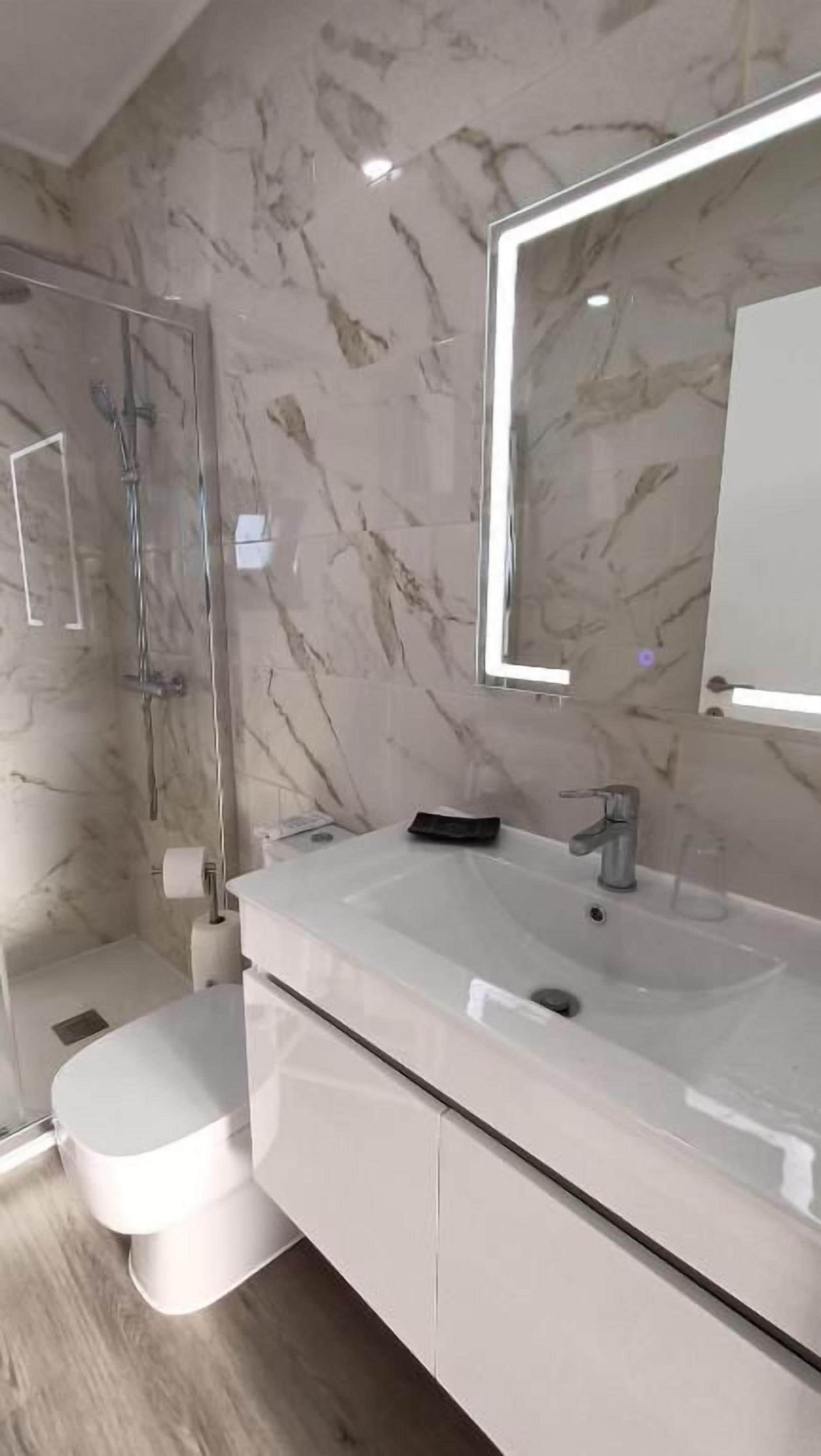 A modern bathroom with a marble wall, a glass shower enclosure, a white sink, and a large mirror.