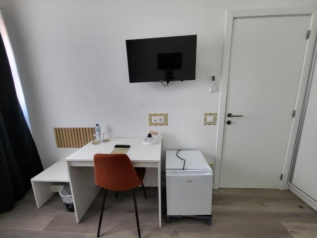 Ein Zimmer mit weißem Schreibtisch, einem an der Wand befestigten Fernseher, einem kleinen weißen Kühlschrank und einer Tür.