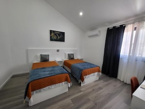A hotel room with two beds, a desk, a chair, a painting on the wall, and a window with curtains.