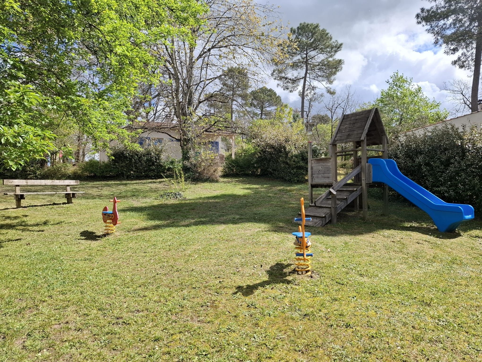 Un terrain de jeux avec un toboggan et des balançoires, entouré d’herbe et d’arbres.