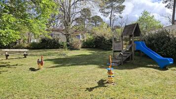 Ein Spielplatz mit Rutsche und Schaukeln, umgeben von Gras und Bäumen.