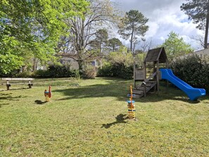 Un terrain de jeux avec un toboggan et des balançoires, entouré d’herbe et d’arbres.