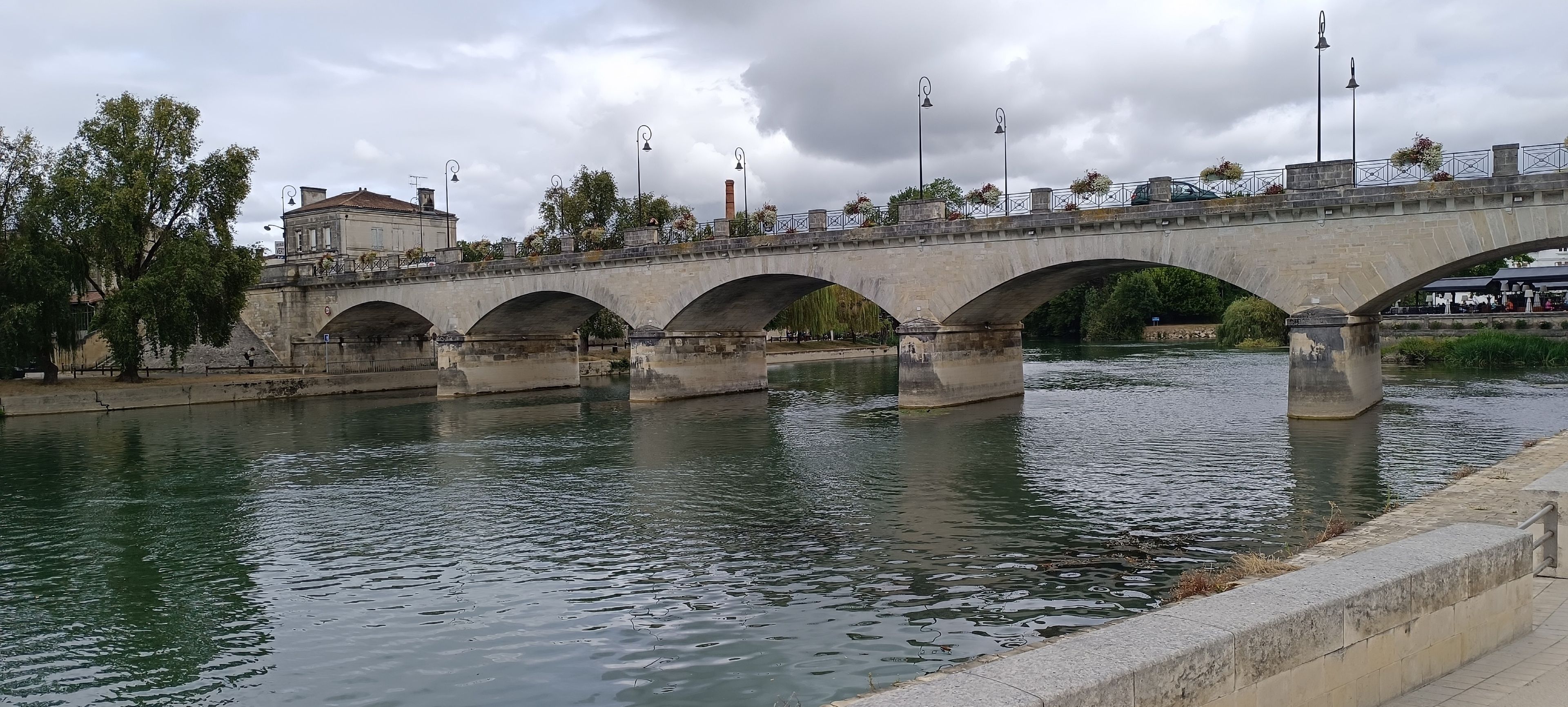Un puente de piedra con varios arcos cruza un río, rodeado de árboles y un edificio de piedra.