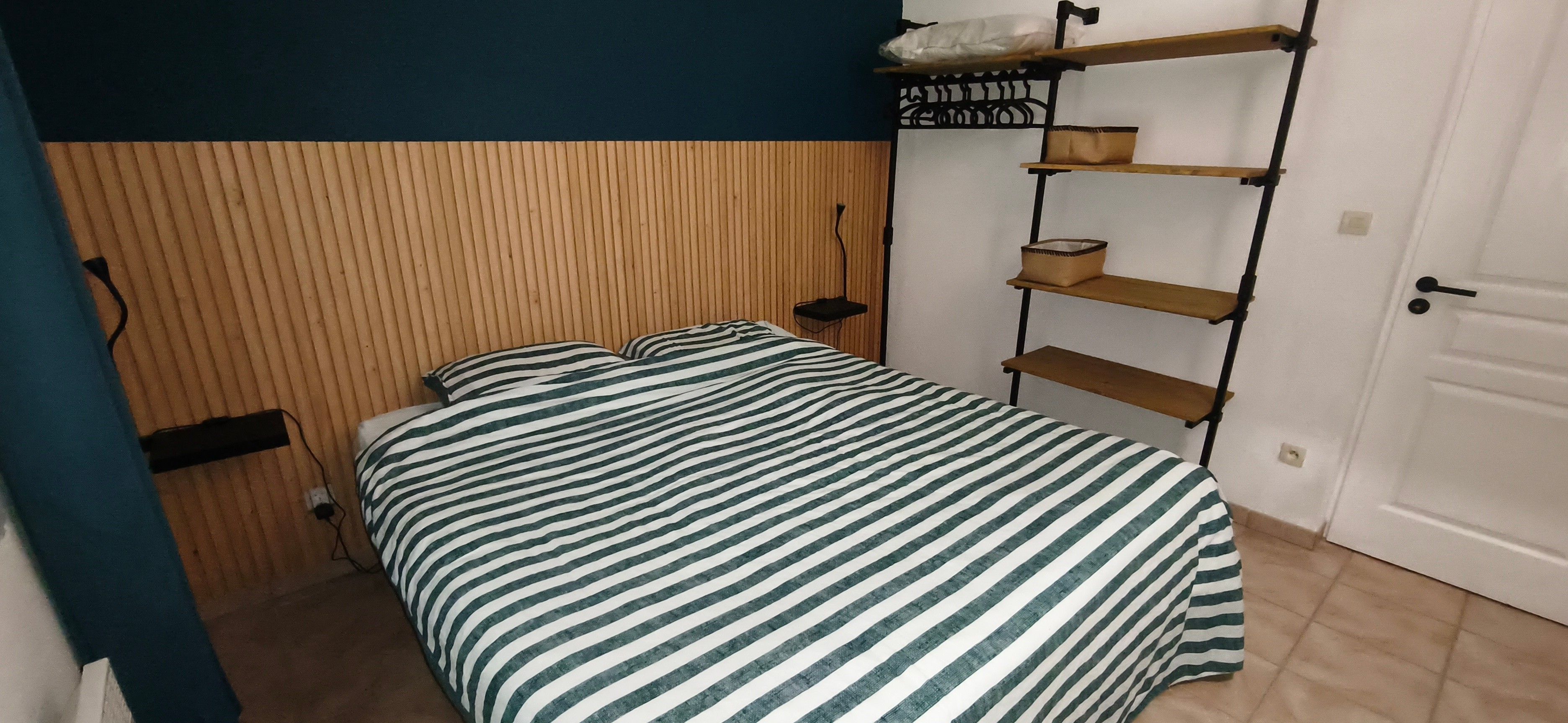 Een slaapkamer met een bed, nachtkastje, een wandplank en een deur.