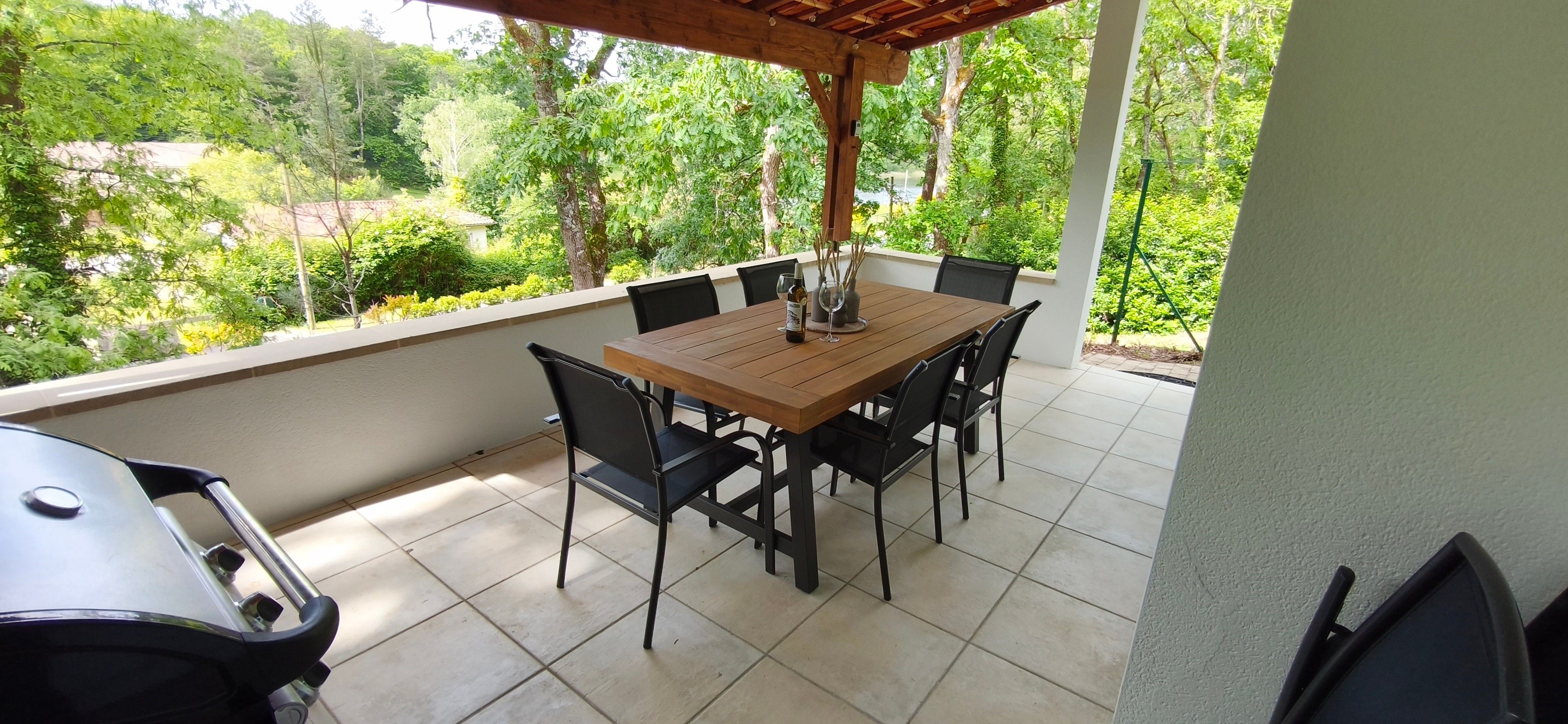 Une terrasse couverte avec une table et des chaises en bois, un barbecue et une vue sur un jardin luxuriant.