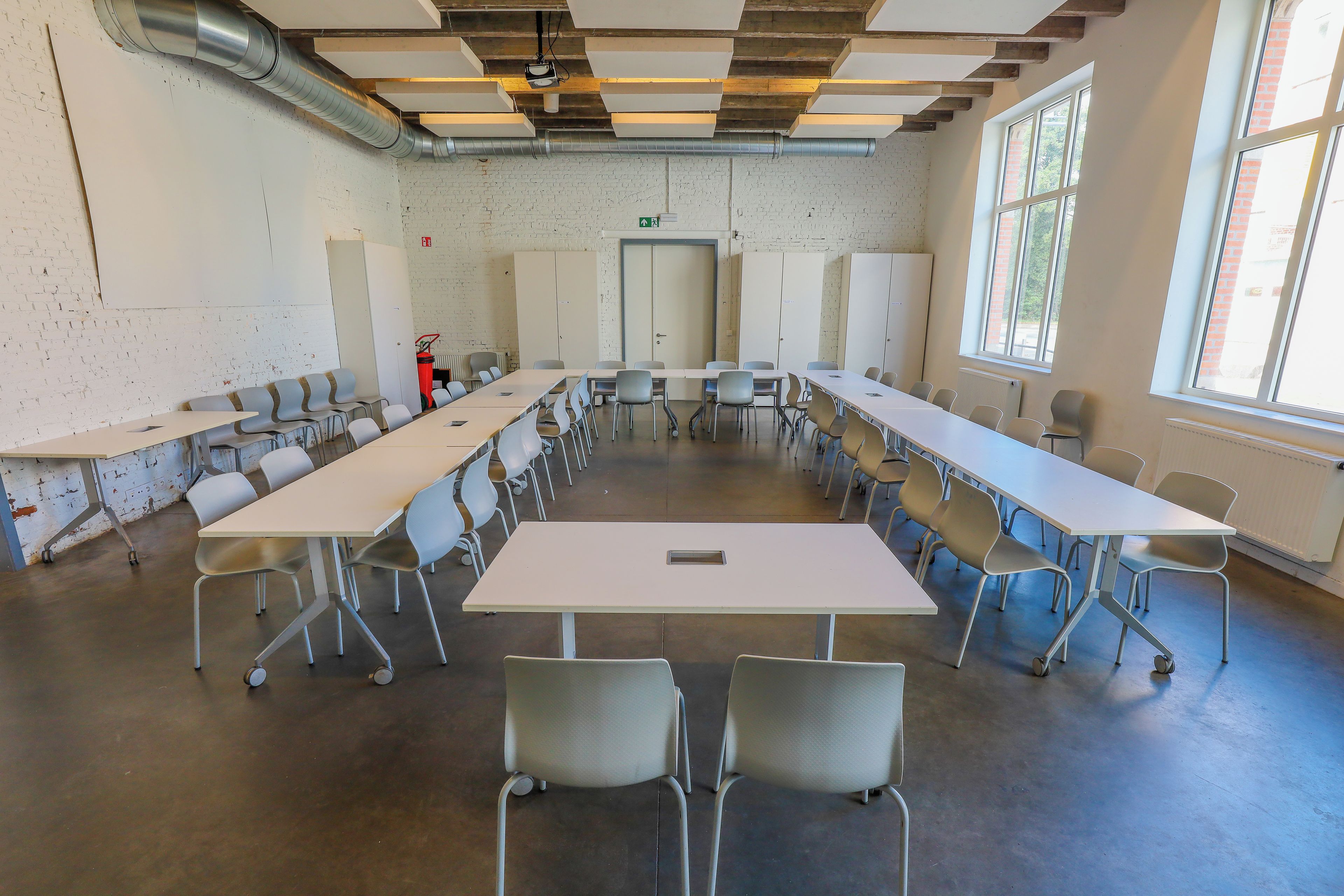 Un salón de conferencias con mesas y sillas blancas, un extintor y una puerta con señal de salida verde.