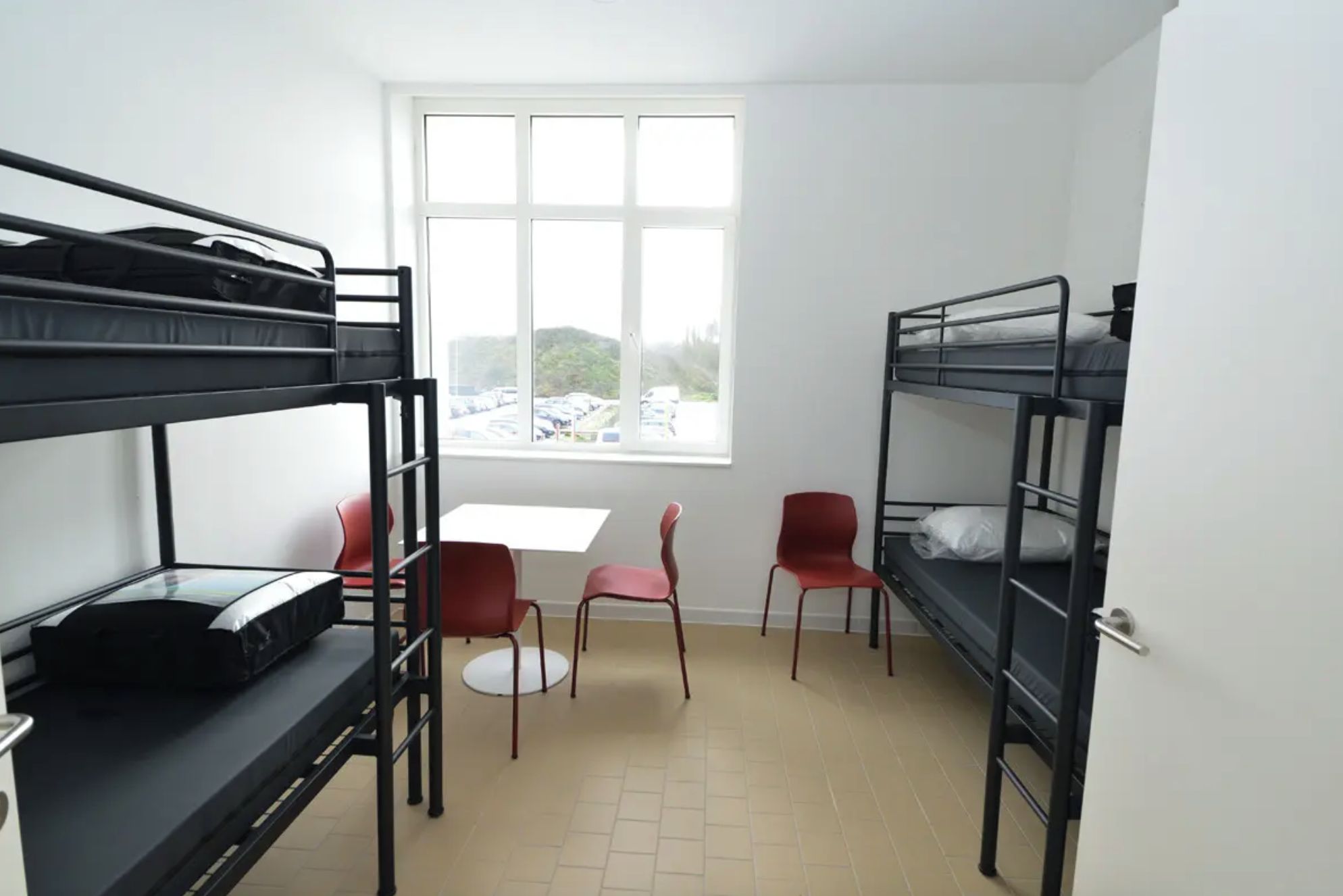 A dormitory room with bunk beds, a small table, and two red chairs.