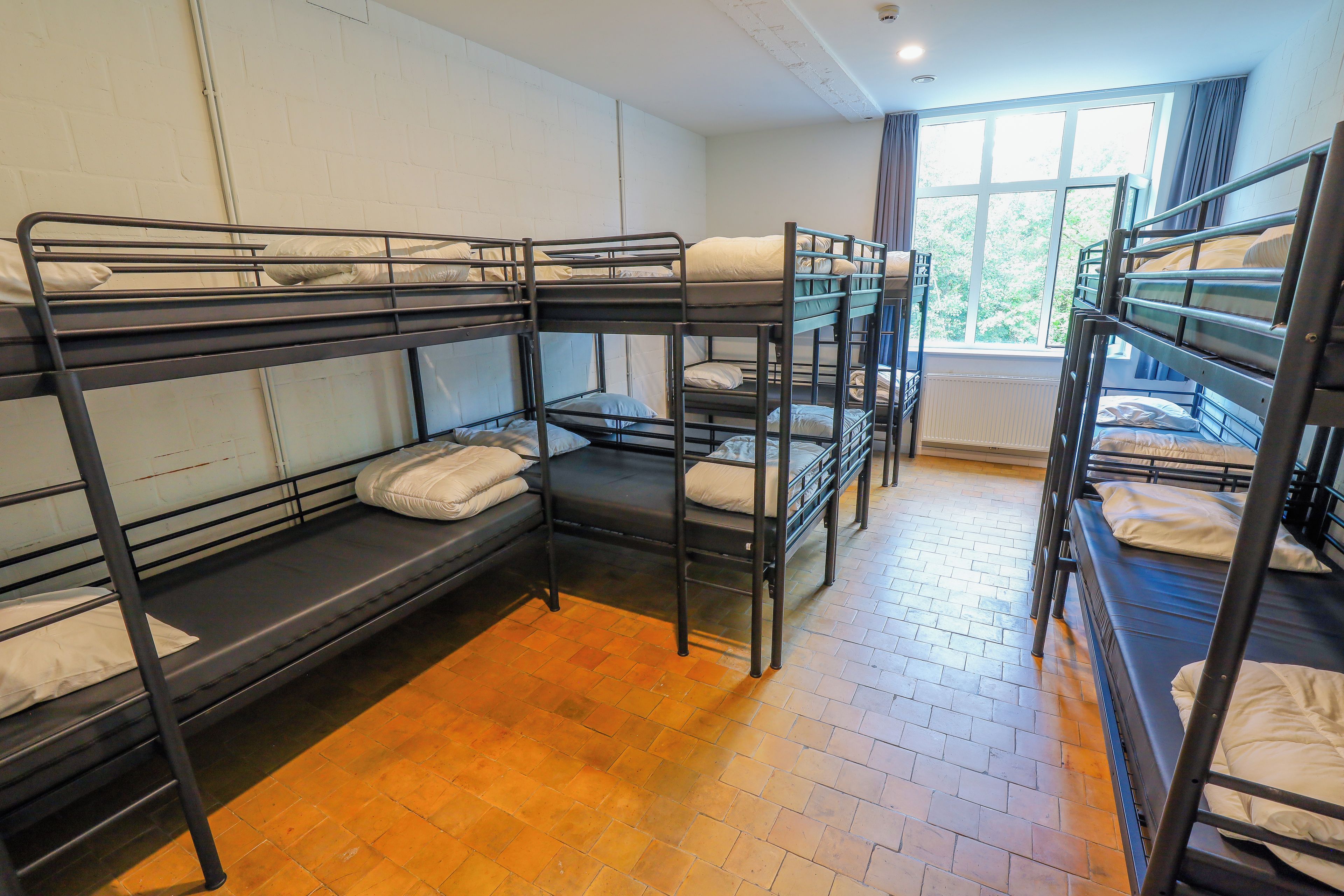 Een slaapzaal met stapelbedden, een raam met gordijnen en een radiator.