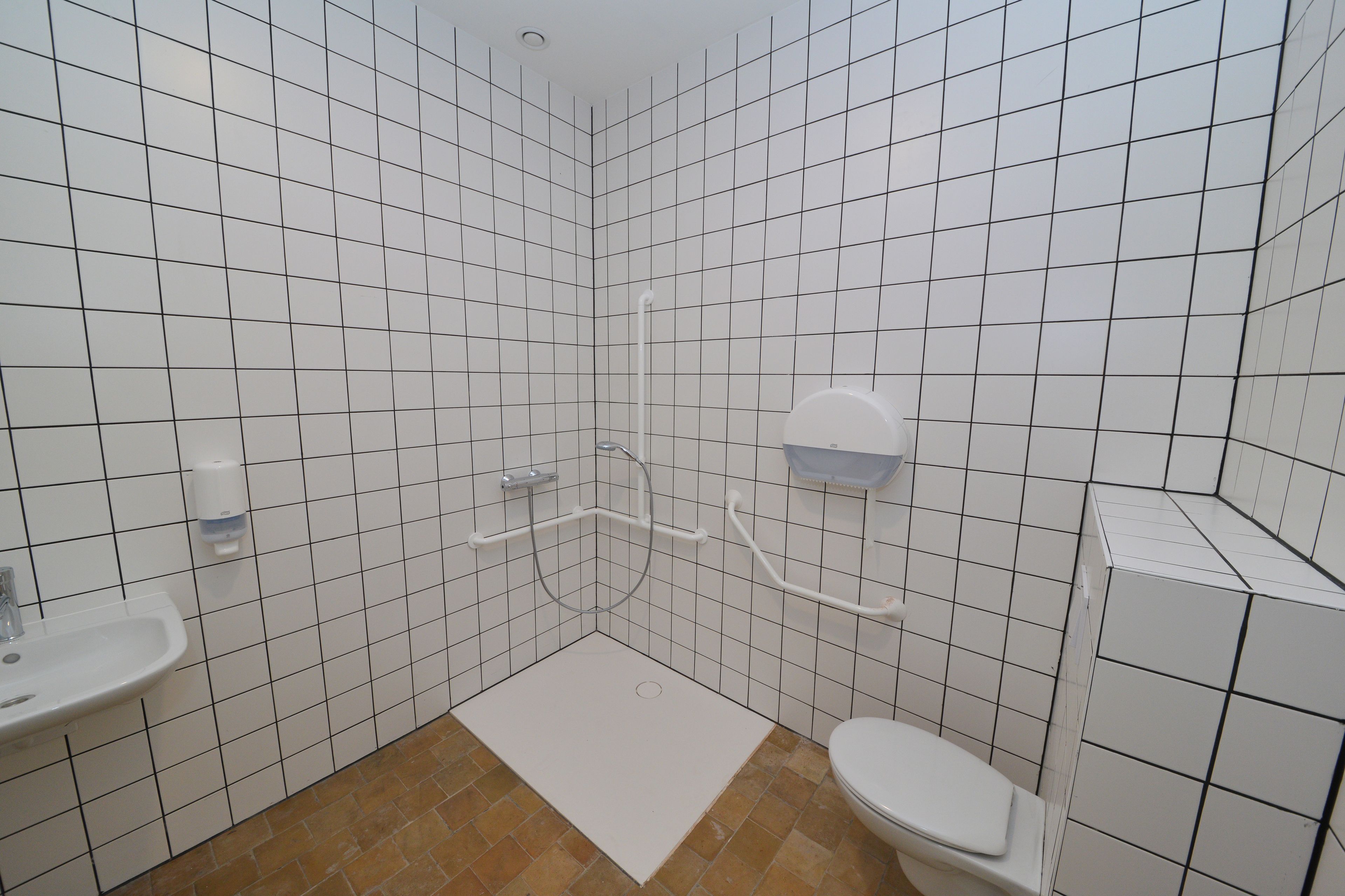 Een badkamer met witte betegelde wanden, een wit wastafelmeubel, een toilet en een douche met een handgreep.