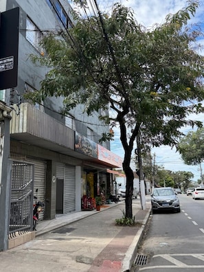 Vista da rua com calçada arborizada, lojas fechadas e carros estacionados.