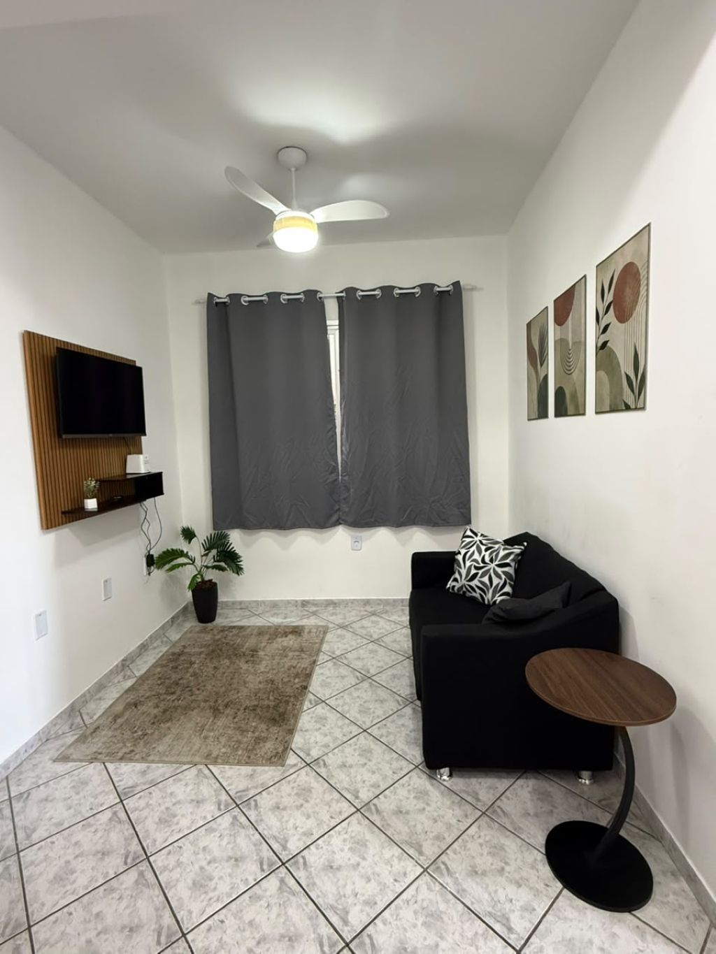 Ein modernes Wohnzimmer mit einer schwarzen Couch, einem kleinen runden Tisch, einem Deckenventilator, einem Flachbildfernseher, einem Blumentopf und Kunstwerken an den Wänden.