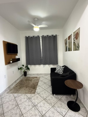 Uma sala de estar moderna com um sofá preto, uma pequena mesa redonda, um ventilador de teto, uma televisão de tela plana, um vaso de planta e obras de arte nas paredes.