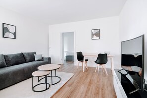 En moderne stue med sofa, to stole, et lille rundt bord, et fjernsyn og et køkken i baggrunden.