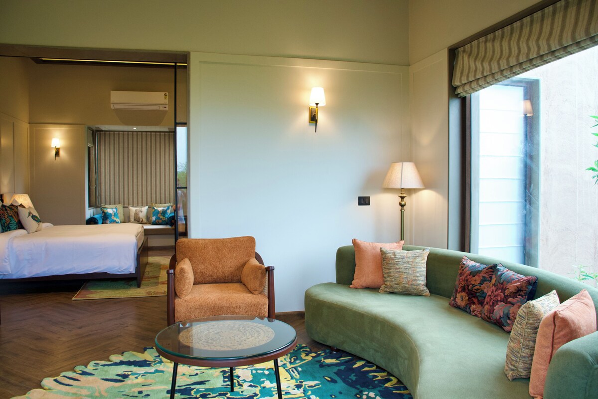 Quarto de hotel com cama, sofá, poltrona, mesa pequena e janela com cortinas.