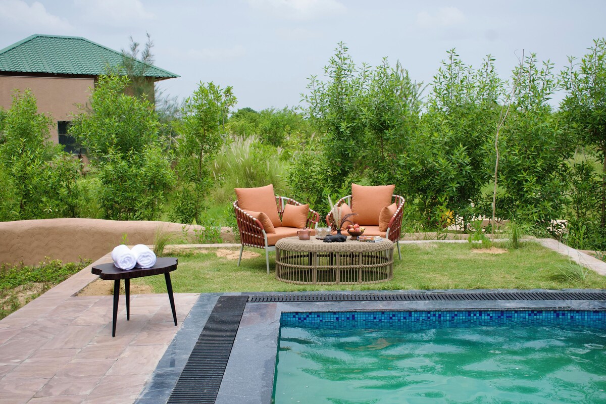 Área de piscina com duas cadeiras de vime, uma mesinha com toalhas e vista para um edifício ao fundo.