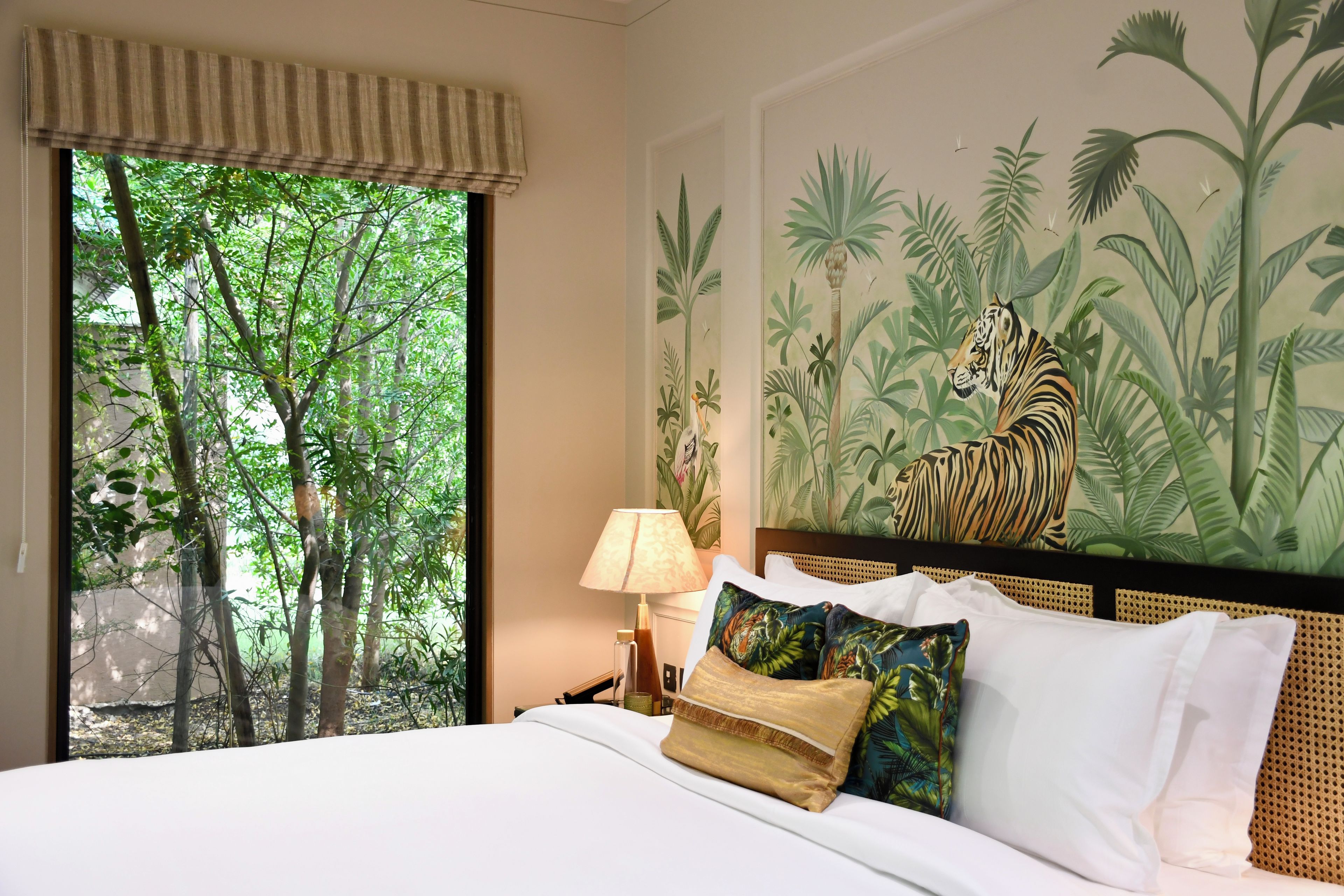 Habitación de hotel con una cama, una mesita de noche, una lámpara, un ventanal con vista a árboles y un mural de tigre en la pared.