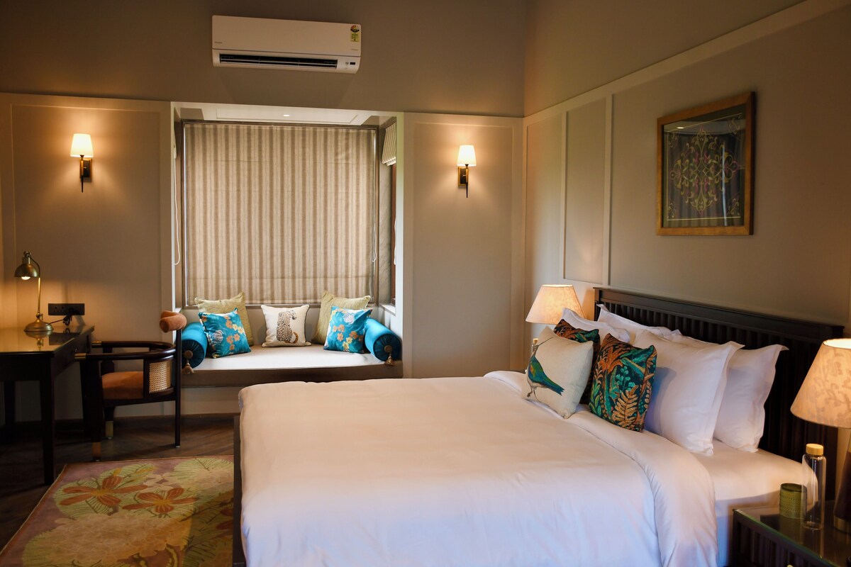 Quarto de hotel com cama, escrivaninha, cadeira, janela com cortinas, ar condicionado fixado na parede e um quadro.