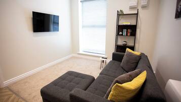 Ein Wohnzimmer mit einem an der Wand montierten Flachbildfernseher, einem grauen Sofa mit Kissen, einem kleinen Beistelltisch und einem Bücherregal mit Büchern und Dekorationsgegenständen.