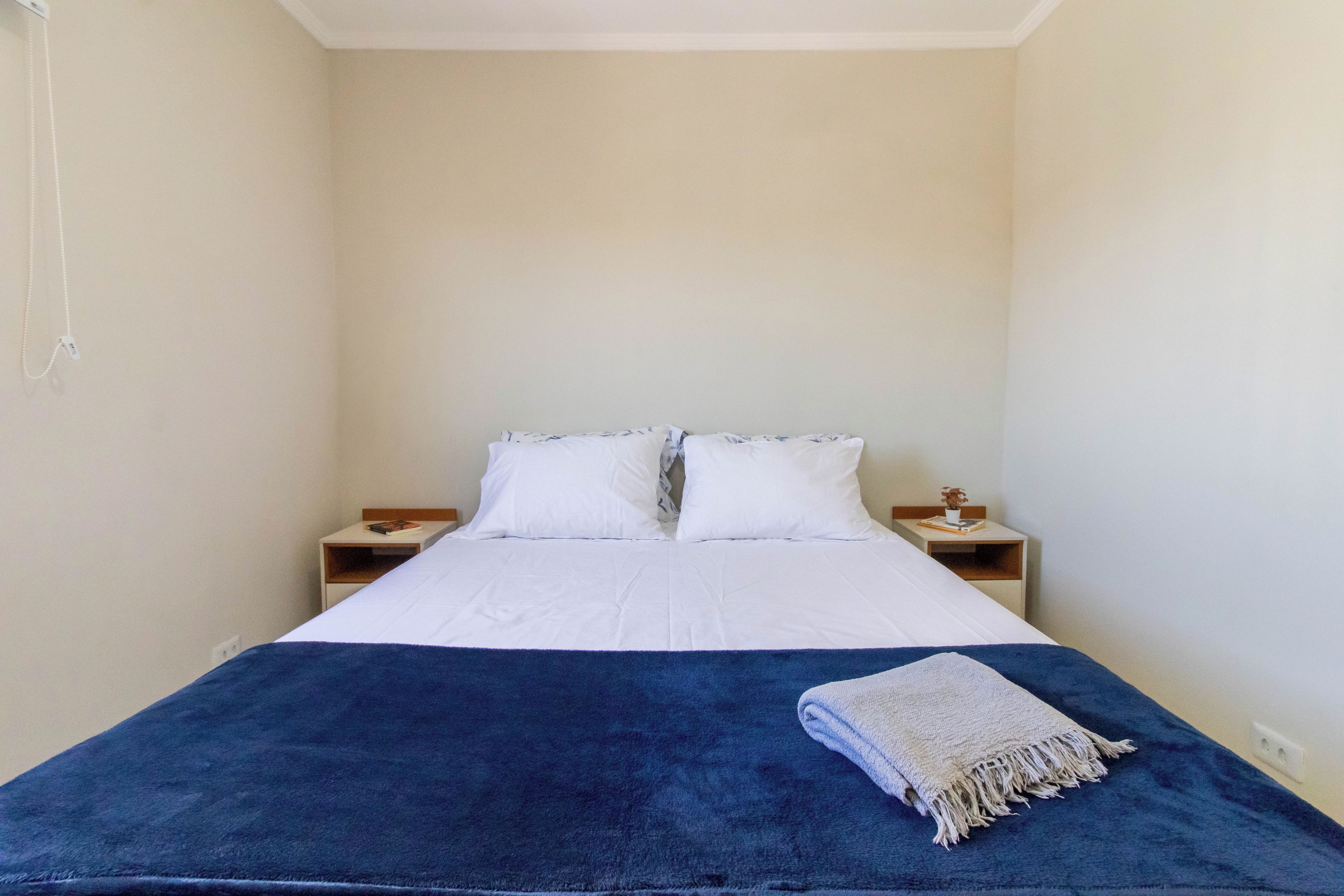 Un letto singolo con lenzuola bianche, due cuscini e una coperta blu. Sono presenti due comodini, uno dei quali con libri e una piccola pianta.