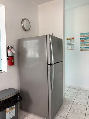 Un réfrigérateur en acier inoxydable dans une cuisine dont le sol est carrelé, avec un extincteur accroché au mur.