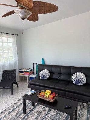 A living room with a black sofa, two decorative pillows, a ceiling fan, a window with curtains, a small table with snacks, and a remote control. - Brisas Del Atlantico House (Luquillo)