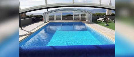 A swimming pool with a glass enclosure, surrounded by a patio area with lounge chairs and a view of the landscape.