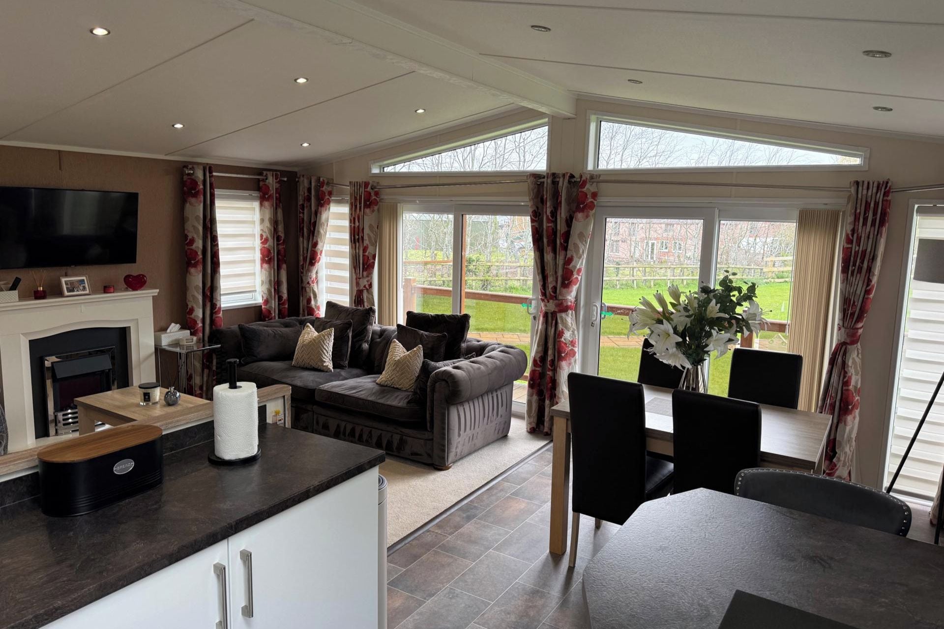 A spacious living area with a fireplace, a sofa set, a dining table with chairs, and large windows offering a view of the outdoors.