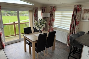 A dining area with a table, chairs, a vase of flowers, and a view of a green lawn through large windows. - Cabin Sleeps 4 With Parking and Balcony (Chester)