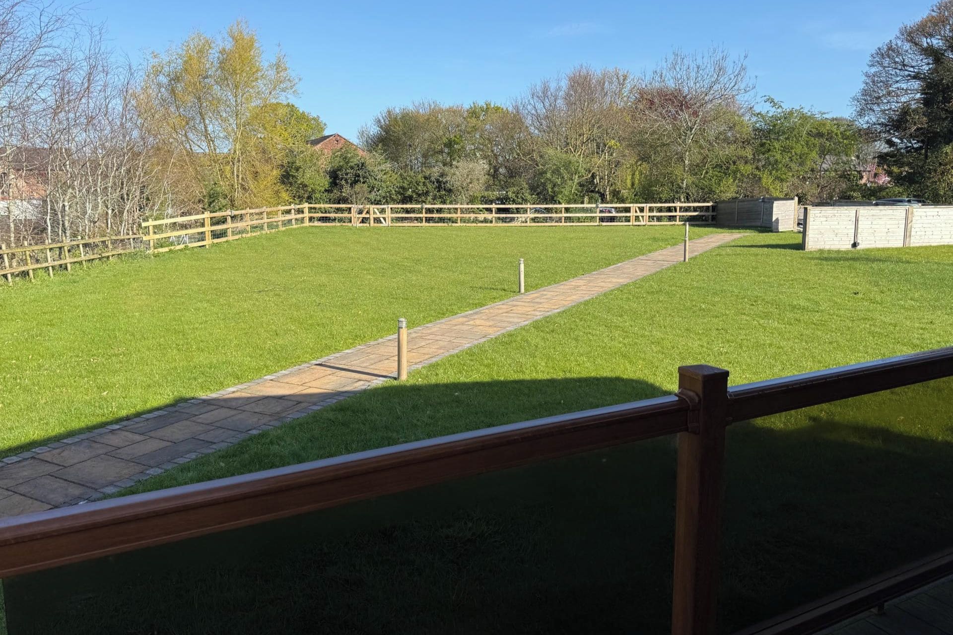 A grassy outdoor area with a paved walkway, surrounded by trees and a wooden fence.