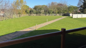 A grassy outdoor area with a paved walkway, surrounded by trees and a wooden fence.