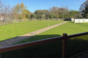 A grassy outdoor area with a paved walkway, surrounded by trees and a wooden fence.