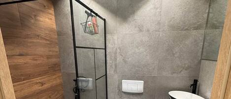 A modern bathroom with a glass shower enclosure, a white toilet, a sink with a black countertop, and grey tiled walls.