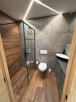 A modern bathroom with a glass shower enclosure, a white toilet, a sink with a black countertop, and grey tiled walls. - defaultName (Besançon)