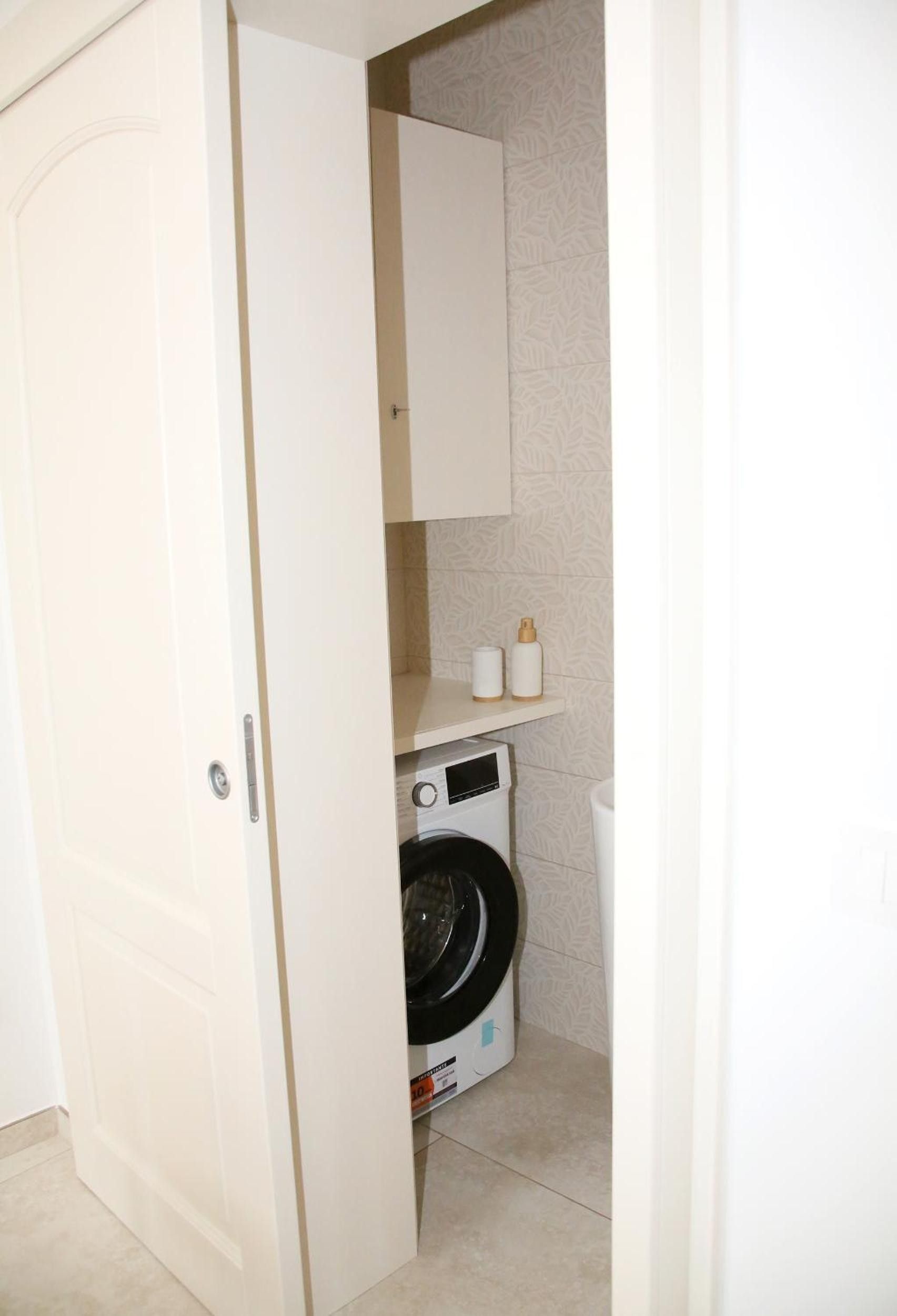 A compact laundry room with a washing machine, a small shelf with toiletries, and a door.