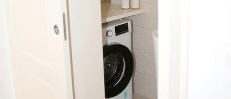 A compact laundry room with a washing machine, a small shelf with toiletries, and a door.