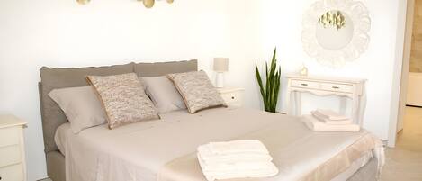 Una cama bien hecha con una colcha blanca y almohadas, una mesita de noche con lámpara y un espejo con marco ornamentado.