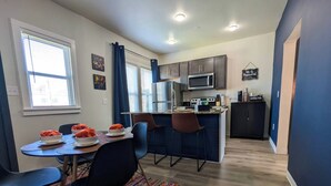 A compact kitchen with modern appliances, a dining area with chairs and a table set for a meal, and a colorful rug on the floor. - Free Parking | Fast Wi-Fi | DT (Detroit)