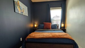 A bedroom with a bed, two bedside lamps, a window with curtains, and a painting on the wall. - Free Parking | Fast Wi-Fi | DT (Detroit)