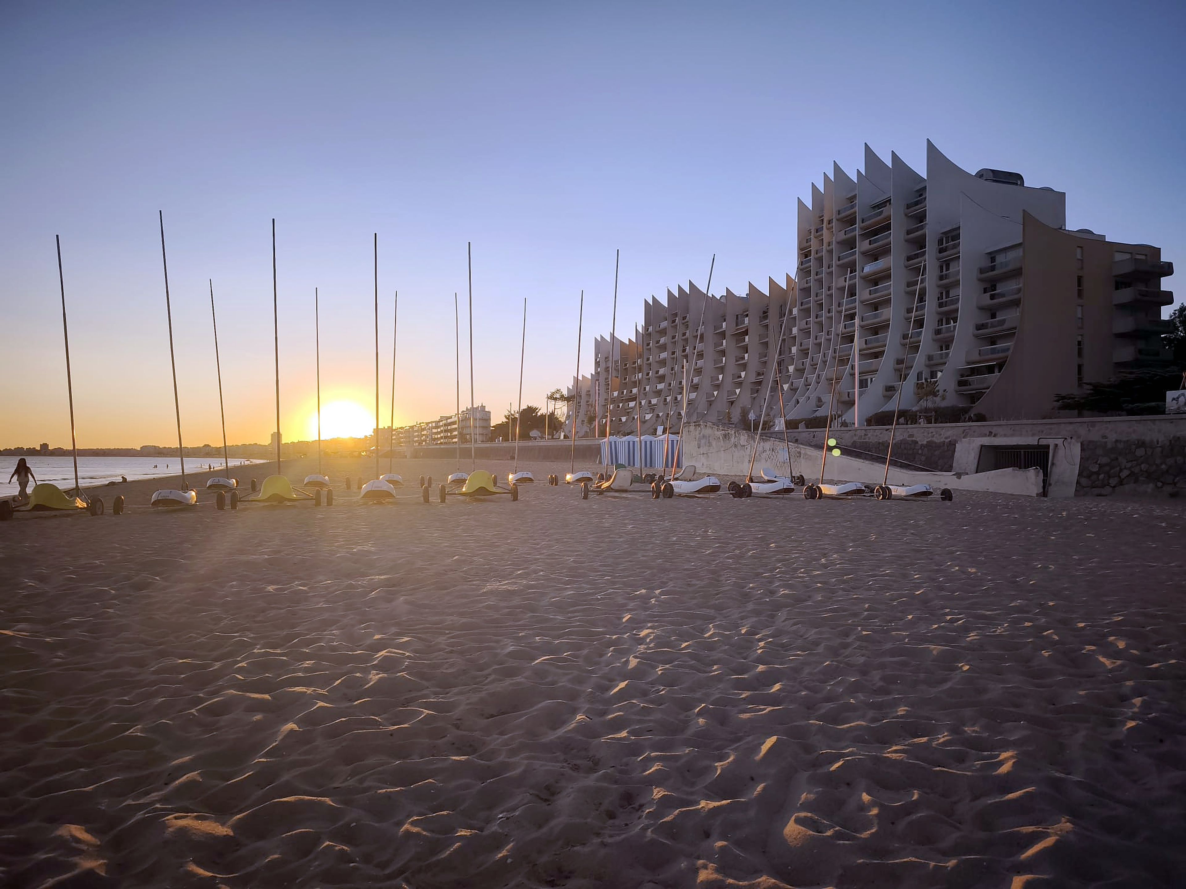 A beach with a row of white beach chairs and umbrellas, a modern building with balconies, and the sun setting in the background.